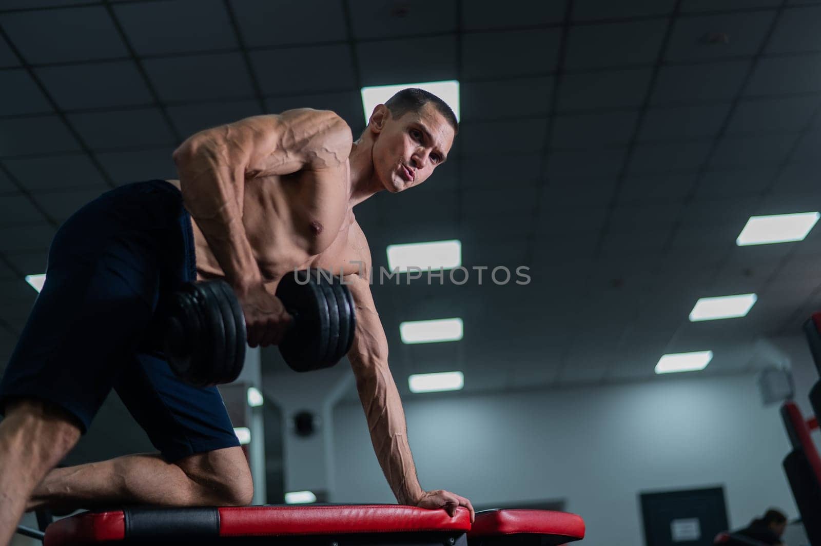 Shirtless man doing dumbbell row to waist on bench at gym