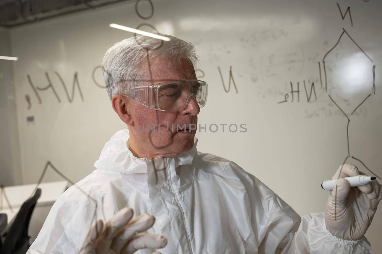 An elderly Caucasian male chemist in a protective suit writes on glass