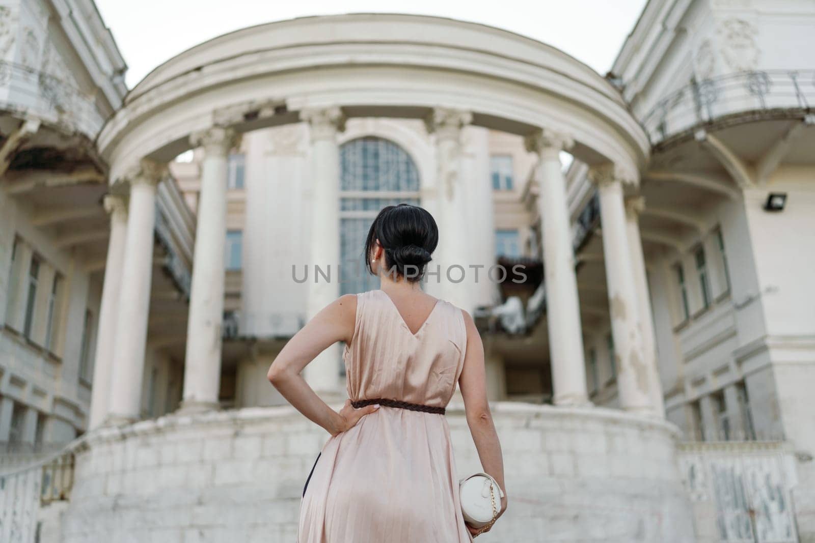A woman in a pink dress stands in front of a large building. She is looking at the camera with a serious expression. The building is old and has a grand appearance. The woman's dress is elegant