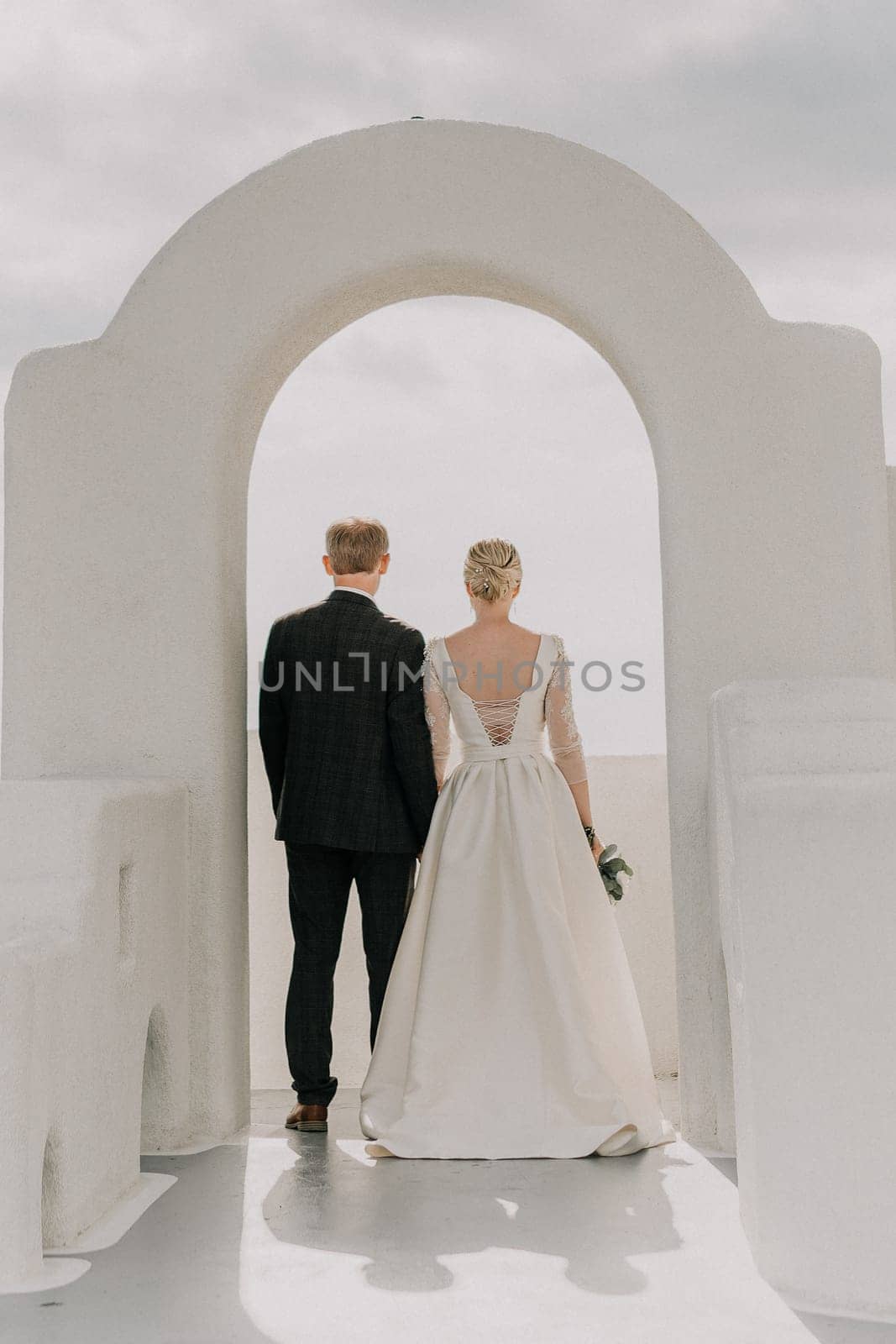 A bride and groom are walking through a white archway on a sunny day. The bride is wearing a white dress and the groom is wearing a suit. The archway is decorated with flowers. by Matiunina