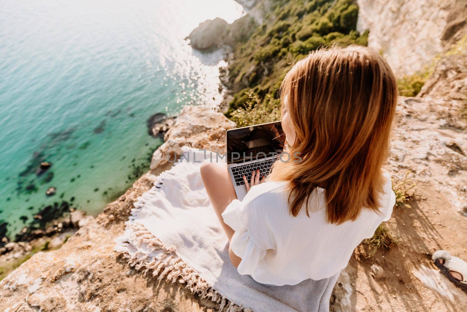 A woman is sitting on a rock overlooking the ocean with a laptop in front of her. She is enjoying the view and the peacefulness of the location