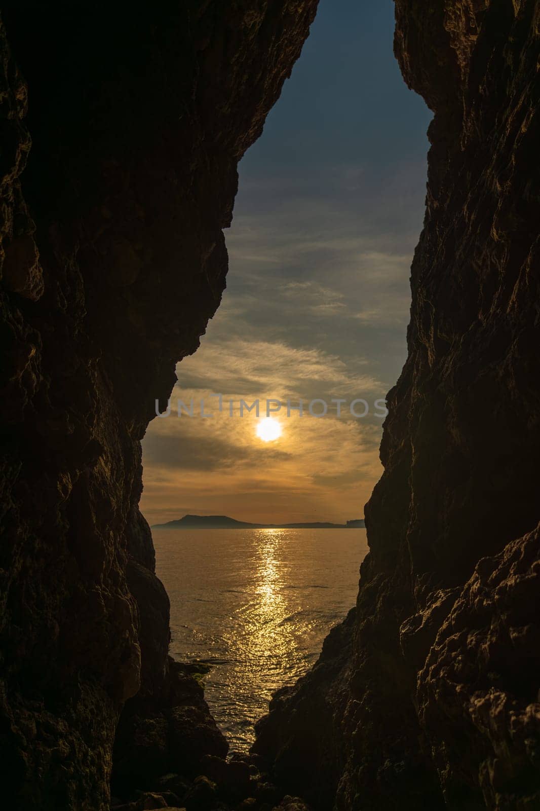 A large rock formation with a small opening in it. The sun is setting and the water is calm