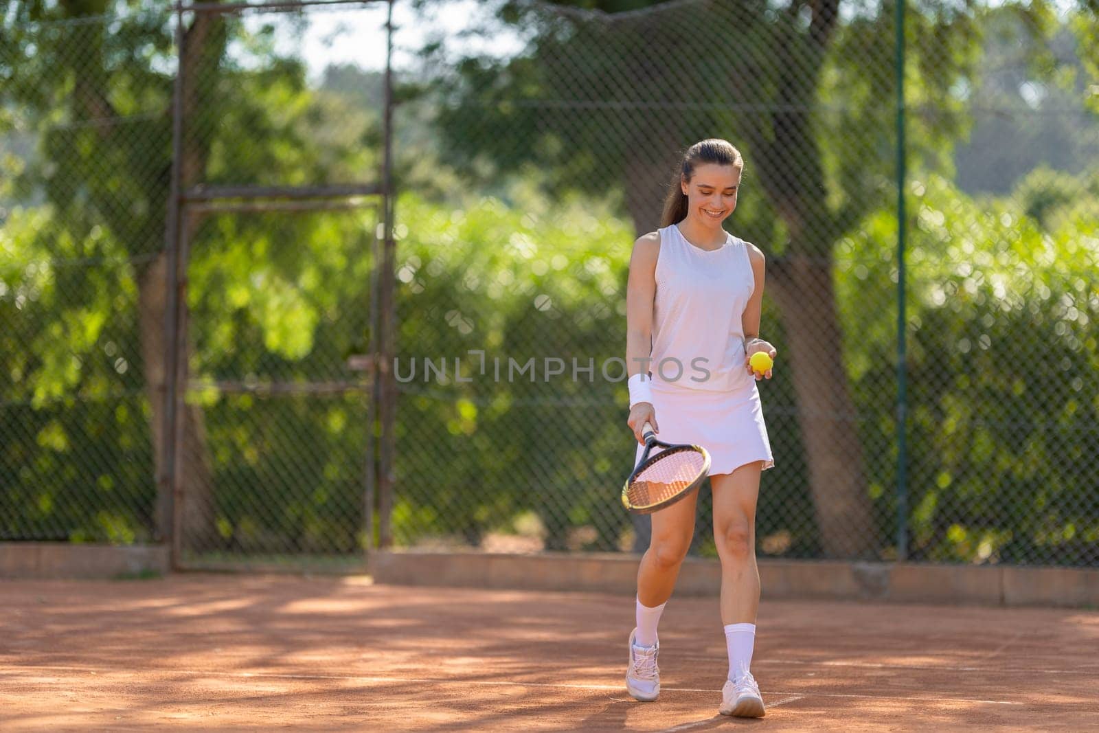 A woman is walking on a tennis court holding a tennis racket and a tennis ball. She is smiling and seems to be enjoying her time