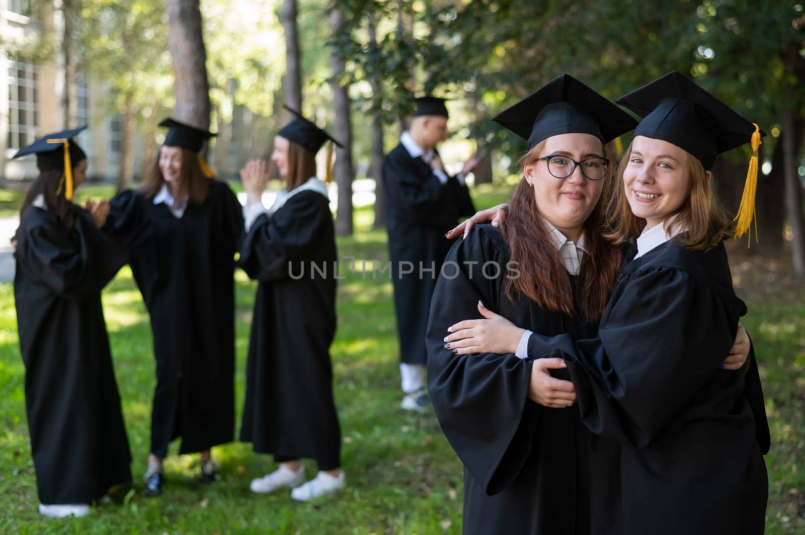 Group of happy students in graduation gowns outdoors. In the foreground, two young girls congratulate each other and embrace