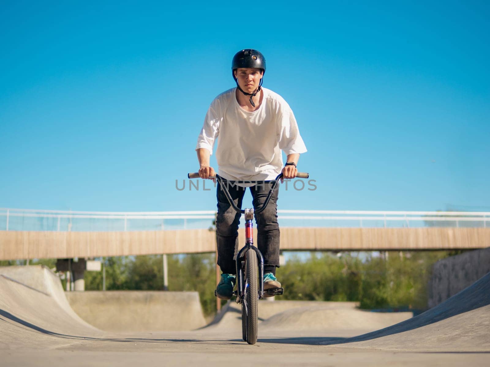 Young BMX bicycle rider having fun and rides in concrete skatepark outdoors. Skilled BMX freestyler in protective helmet posing in quarter pipe in ramp park. Cheerful boy rides on BMX, look at camera