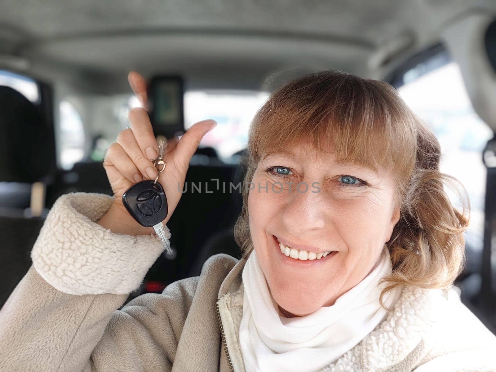 A woman with a bright smile proudly holds up a car key fob while sitting inside her car on chilly winter morning