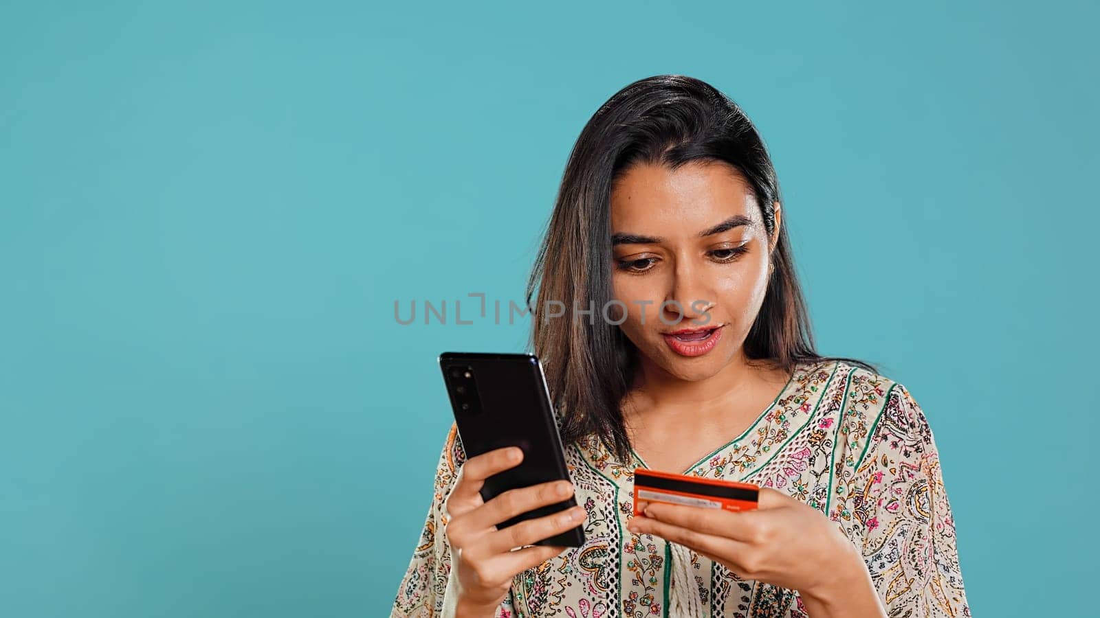 Indian woman excited to purchasing stuff, adding payment method on mobile phone website, isolated over studio background. Ecstatic client does internet shopping, typing debit card info, camera B