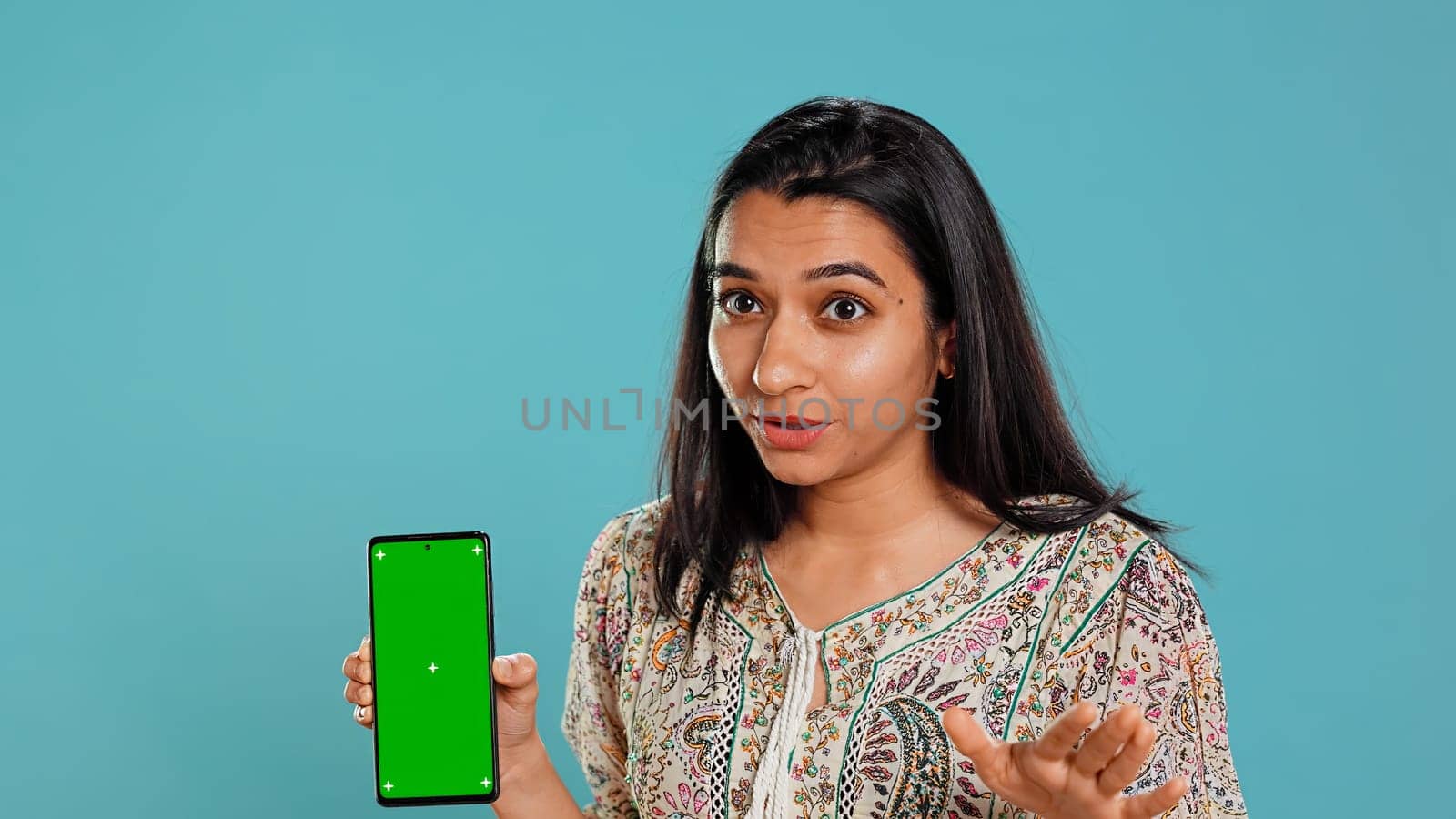 Woman presenting green screen mobile phone, isolated over studio background. Cheerful indian person holding copy space chroma key smartphone used for advertising brands, camera B