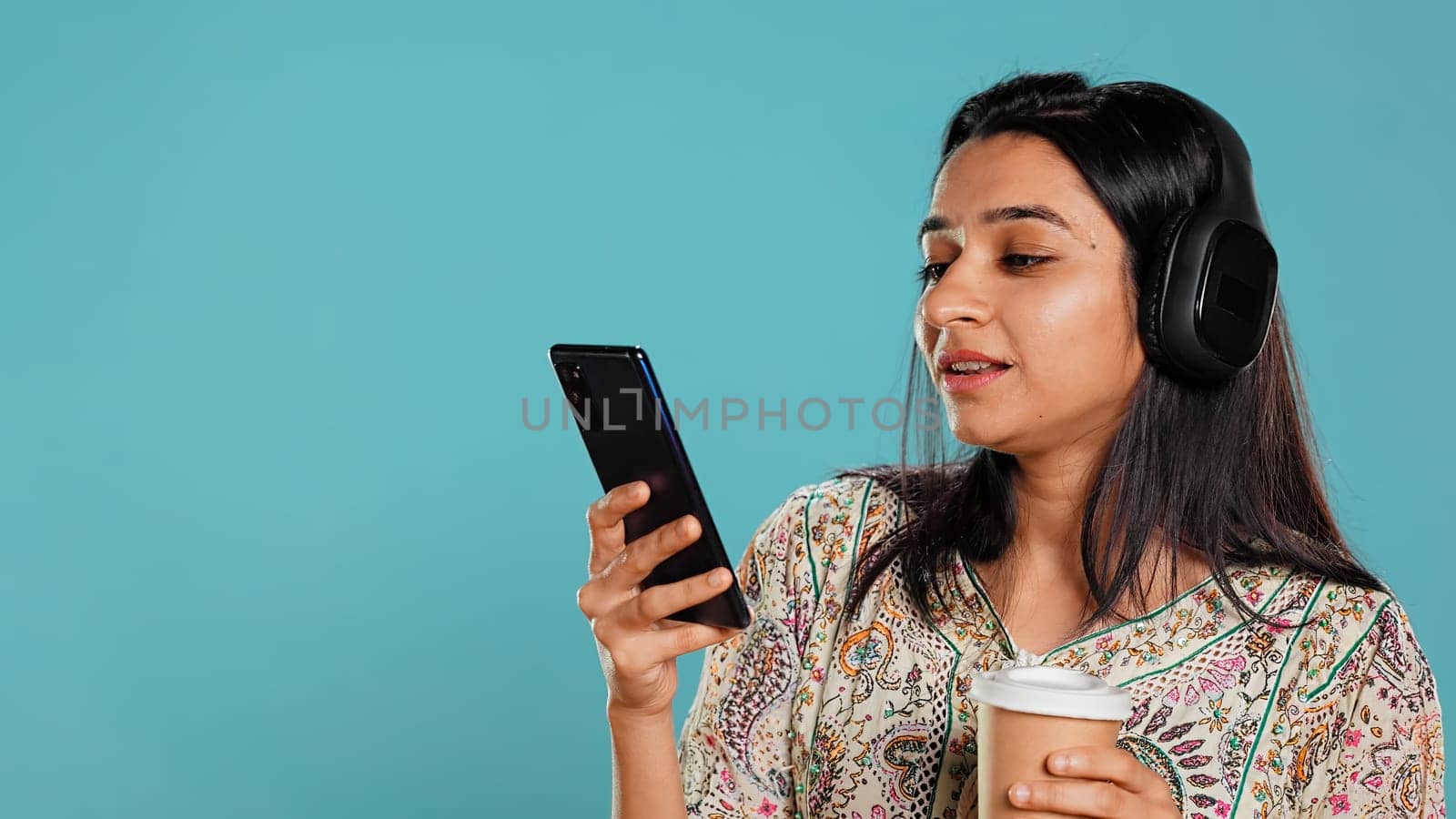 Joyous woman listening music, singing lyrics from phone screen, enjoying beverage. Cheerful person wearing headphones, listening songs, head bopping and holding coffee cup, studio background, camera B