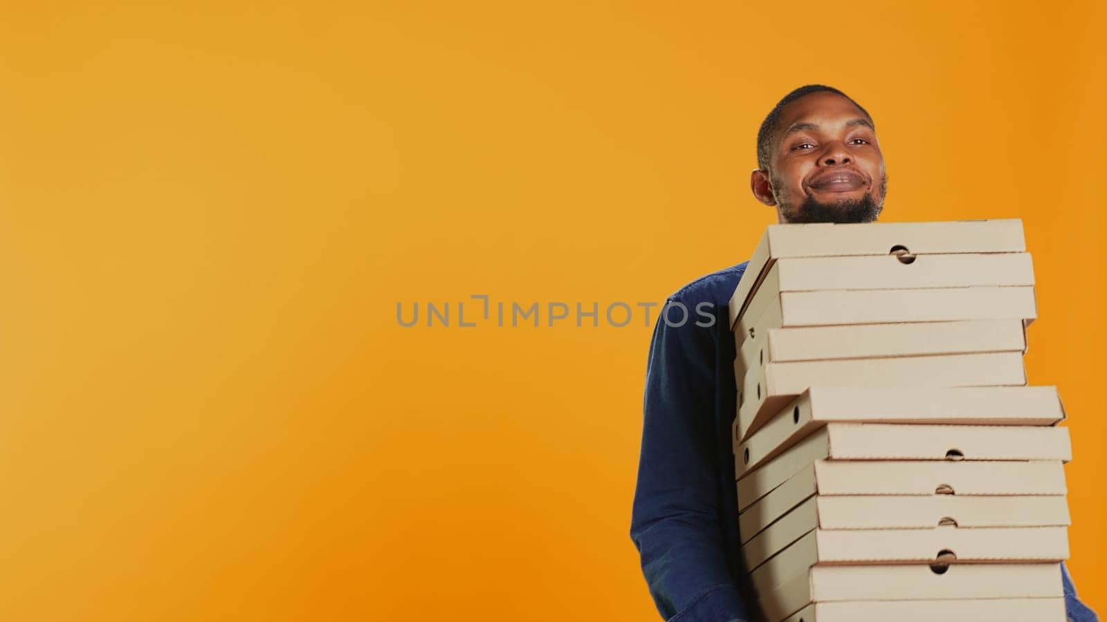 Confident male courier carrying big pile of pizza boxes to deliver fast food order to clients, takeaway service in studio. Young smiling deliveryman bringing takeout meal stack. Camera B.