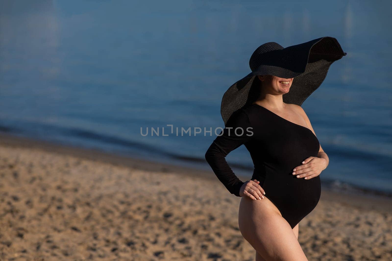 Pregnant woman in a large straw hat and black swimsuit posing on the beach. by mrwed54
