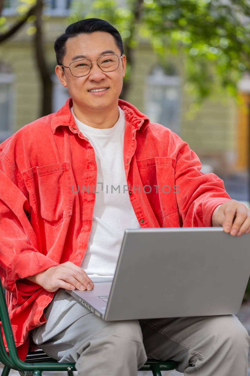 Asian middle-aged man freelancer working online distant job with laptop browsing website chatting sitting outdoors. Chinese guy on city street looking at notebook screen send messages watching movies
