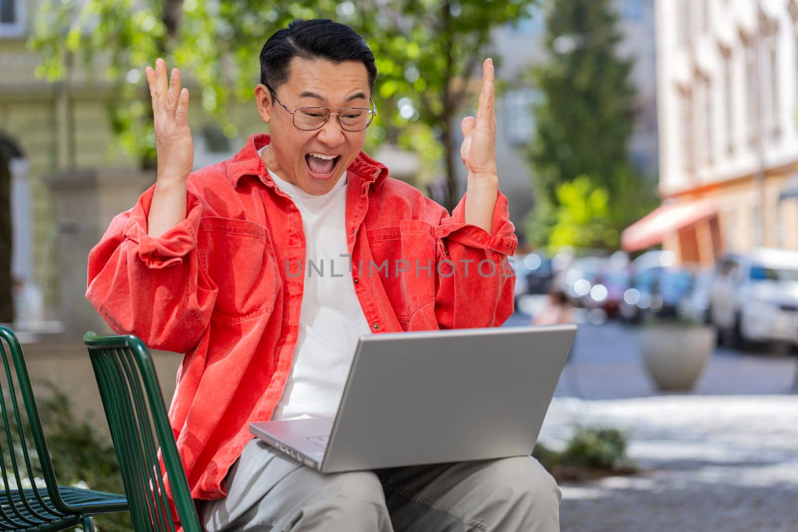 Happy Asian middle-aged man working on laptop screams in triumph win celebrating good message news success game casino lottery money jackpot victory outdoor. Chinese guy on urban city street