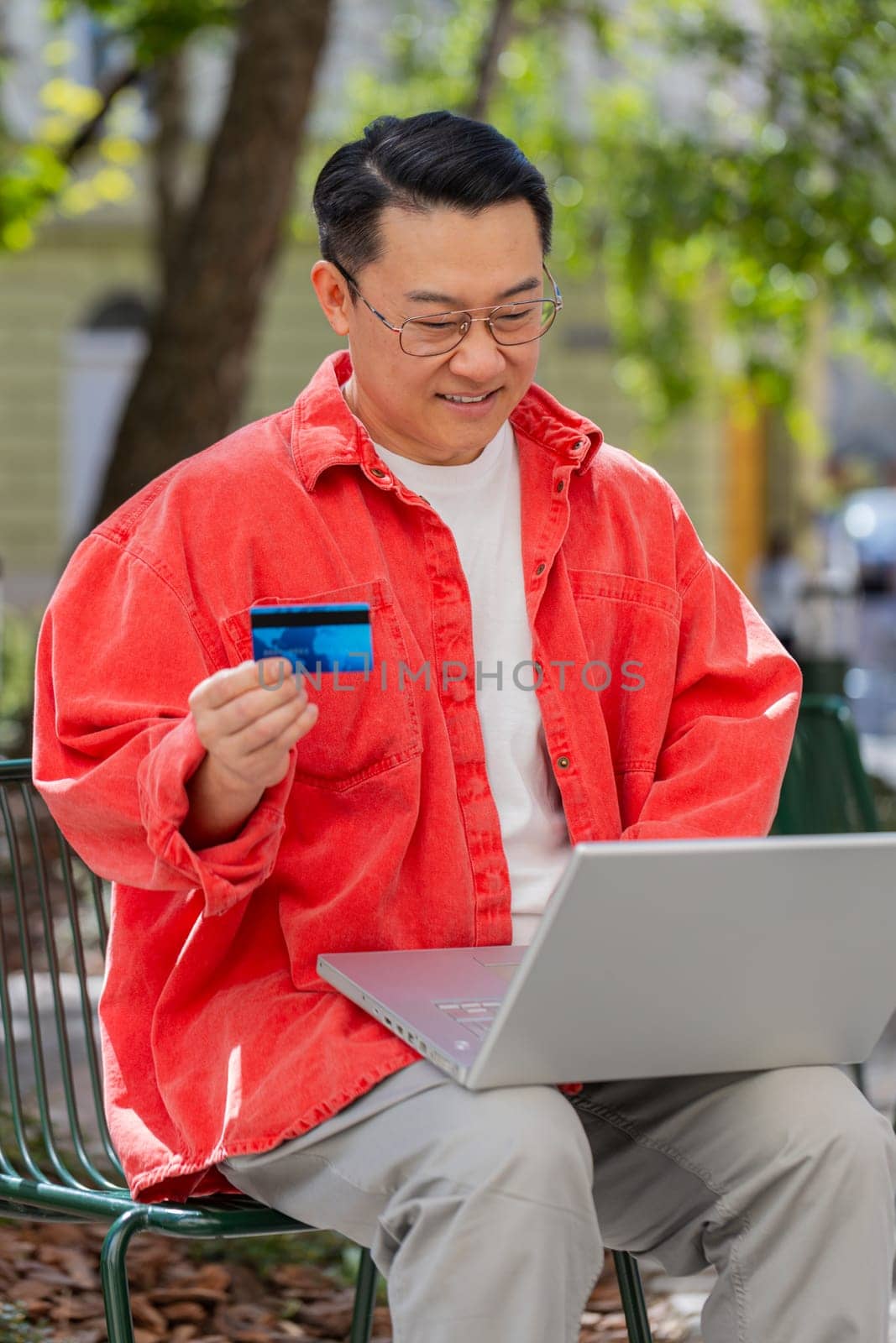 Asian middle-aged man using credit bank plastic card and laptop computer while transferring money purchases online shopping order food delivery on internet outdoors. Chinese guy on urban city street