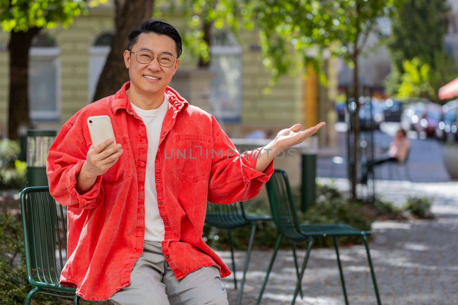 Asian middle-aged man using smartphone typing text messages in social media application online, surfing internet, relaxing, taking a break outdoors. Chinese mature guy sitting on urban city street.