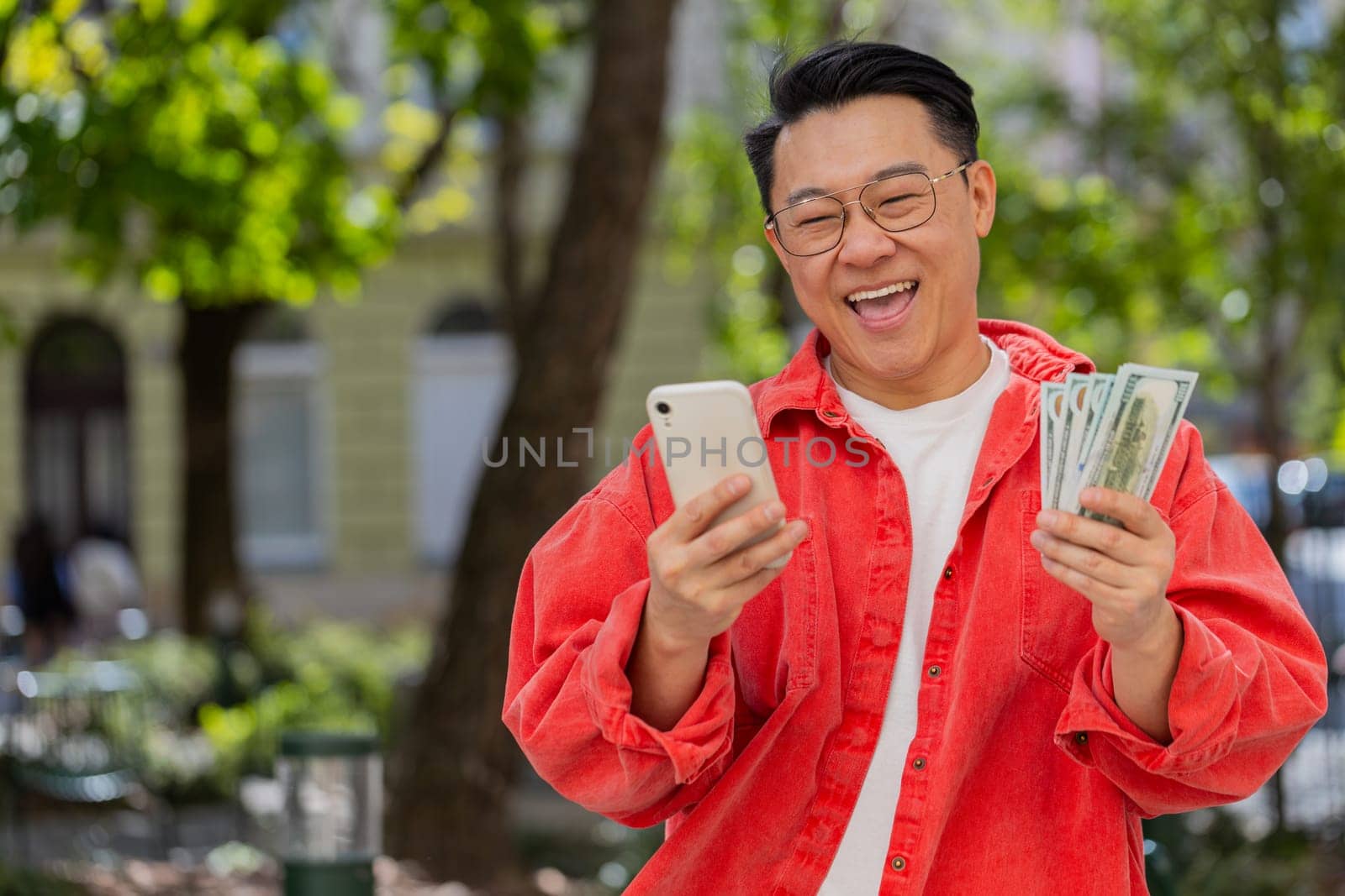 Happy rich Asian middle-aged man counting money dollar cash use smartphone calculator app plans to order gifts or food delivery online booking hotel room outdoors. Chinese guy on urban city street.