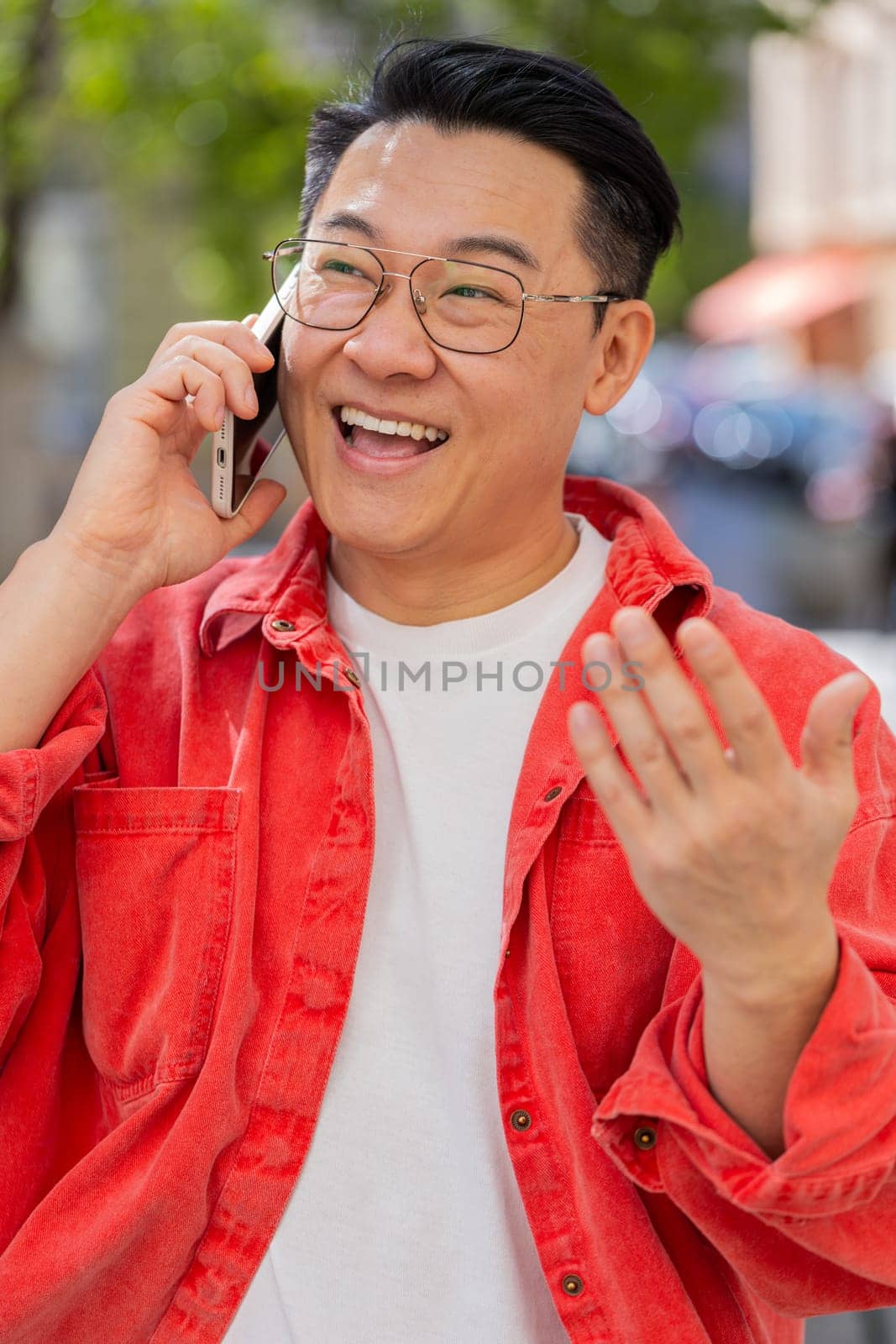 Happy Asian middle-aged man having remote conversation communicate speaking by smartphone outdoors. Japanese guy talking on phone unexpected good news gossip on city street. Town lifestyles. Vertical