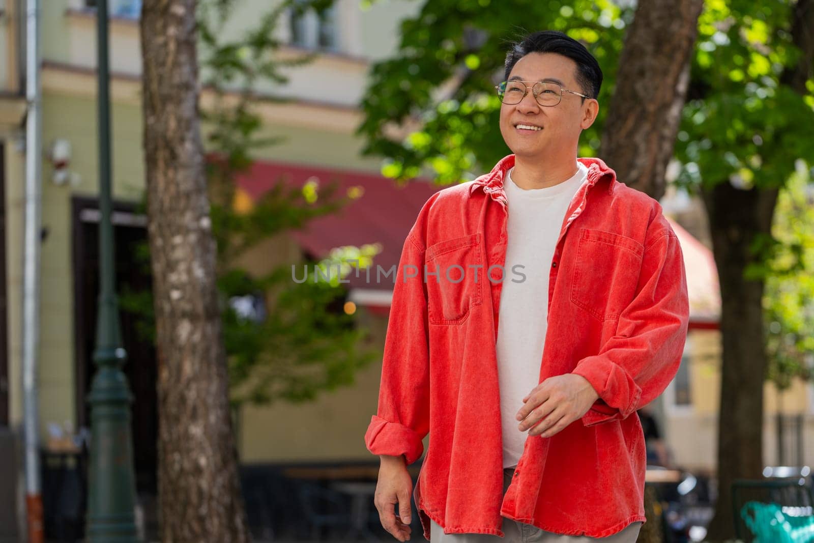Asian middle-aged man tourist walking on urban city street outdoors in summer daytime. Chinese guy traveler smiling having positive good mood enjoying vacation holidays. Town lifestyles. Horizontal