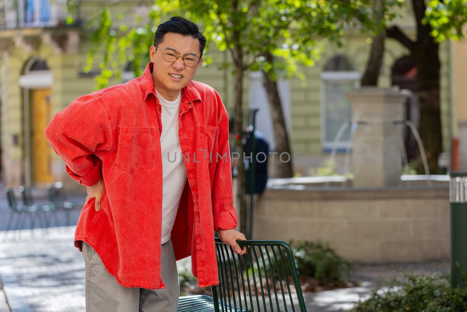 Asian middle-aged man suffering backache massaging sore place lower spine frowning from unbearable spasm torn muscles and tendons kidney stones pinched nerve outdoors. Chinese guy on urban city street