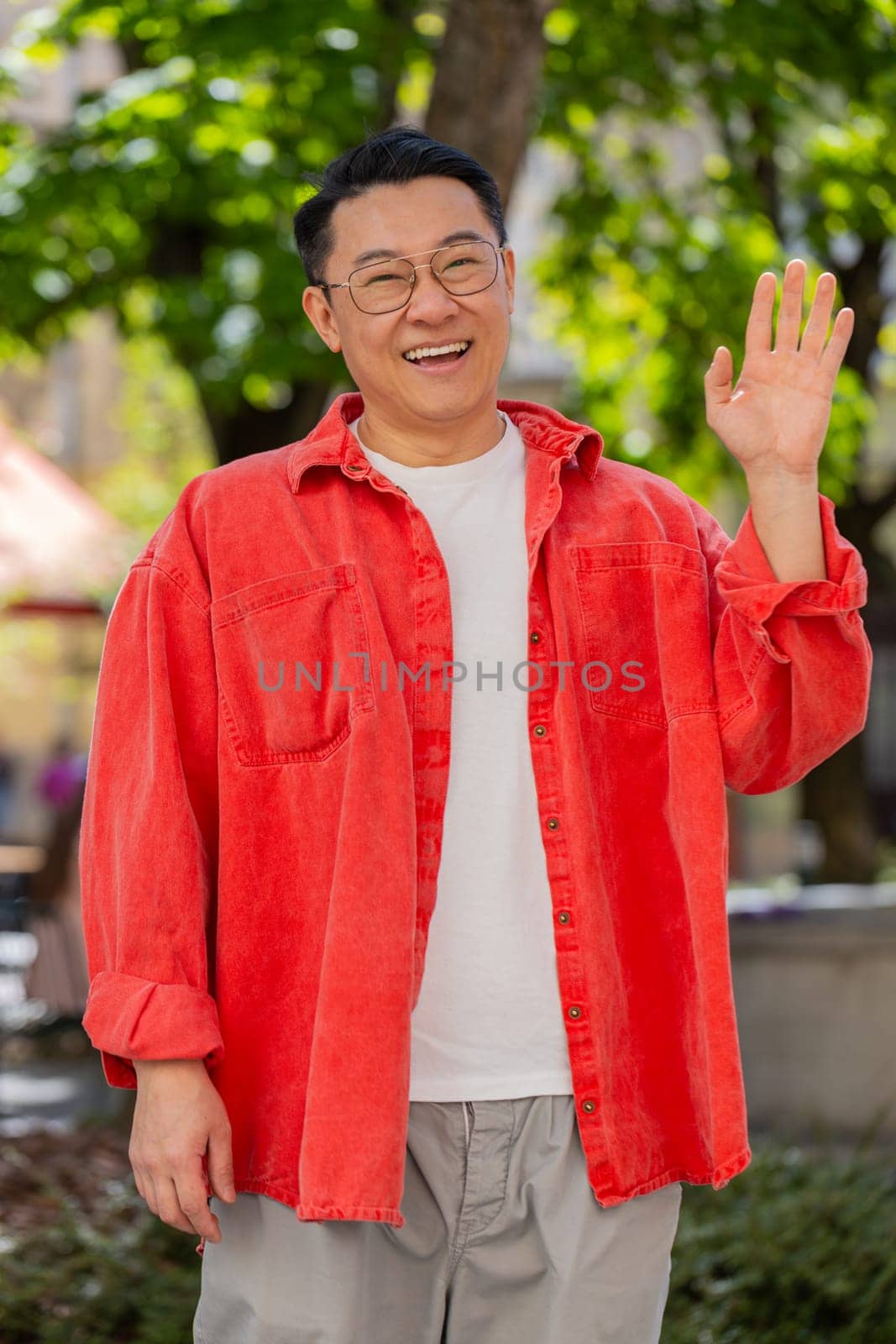 Hello. Asian middle-aged man smiling friendly at camera, waving hands gesturing hi, greeting or goodbye, welcoming with hospitable expression outdoors. Chinese guy on urban city street. Vertical