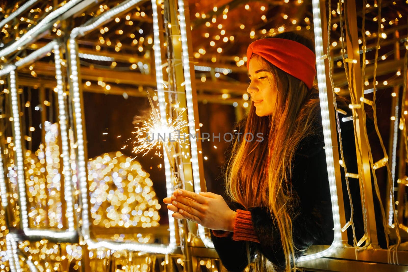 Woman holding sparkler night while celebrating Christmas outside. Dressed in a fur coat and a red headband. Blurred christmas decorations in the background. Selective focus.