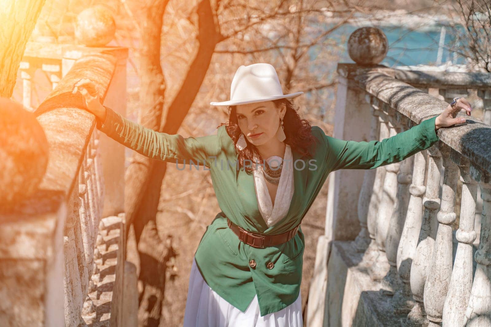 Woman walks around the city, lifestyle. Happy woman in a green jacket, white skirt and hat is sitting on a white fence with balusters overlooking the sea bay and the city