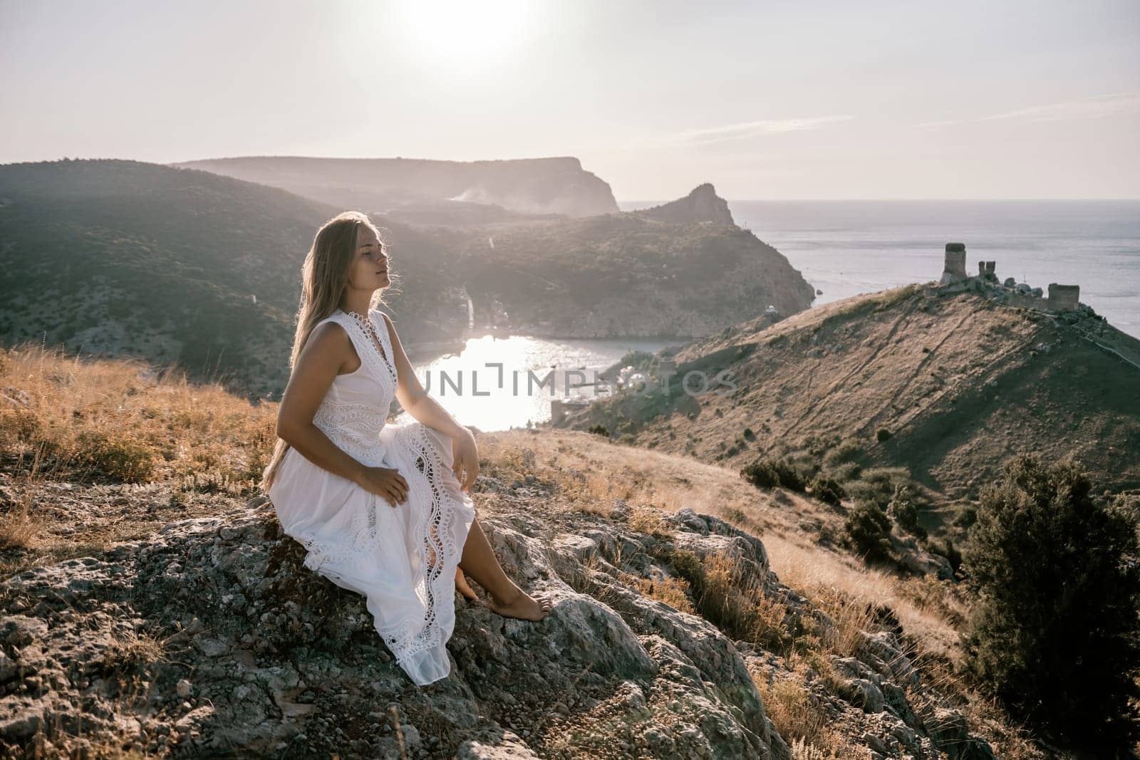 A woman in a white dress sits on a rock overlooking a body of water. The scene is serene and peaceful, with the woman enjoying the view and the calmness of the surroundings