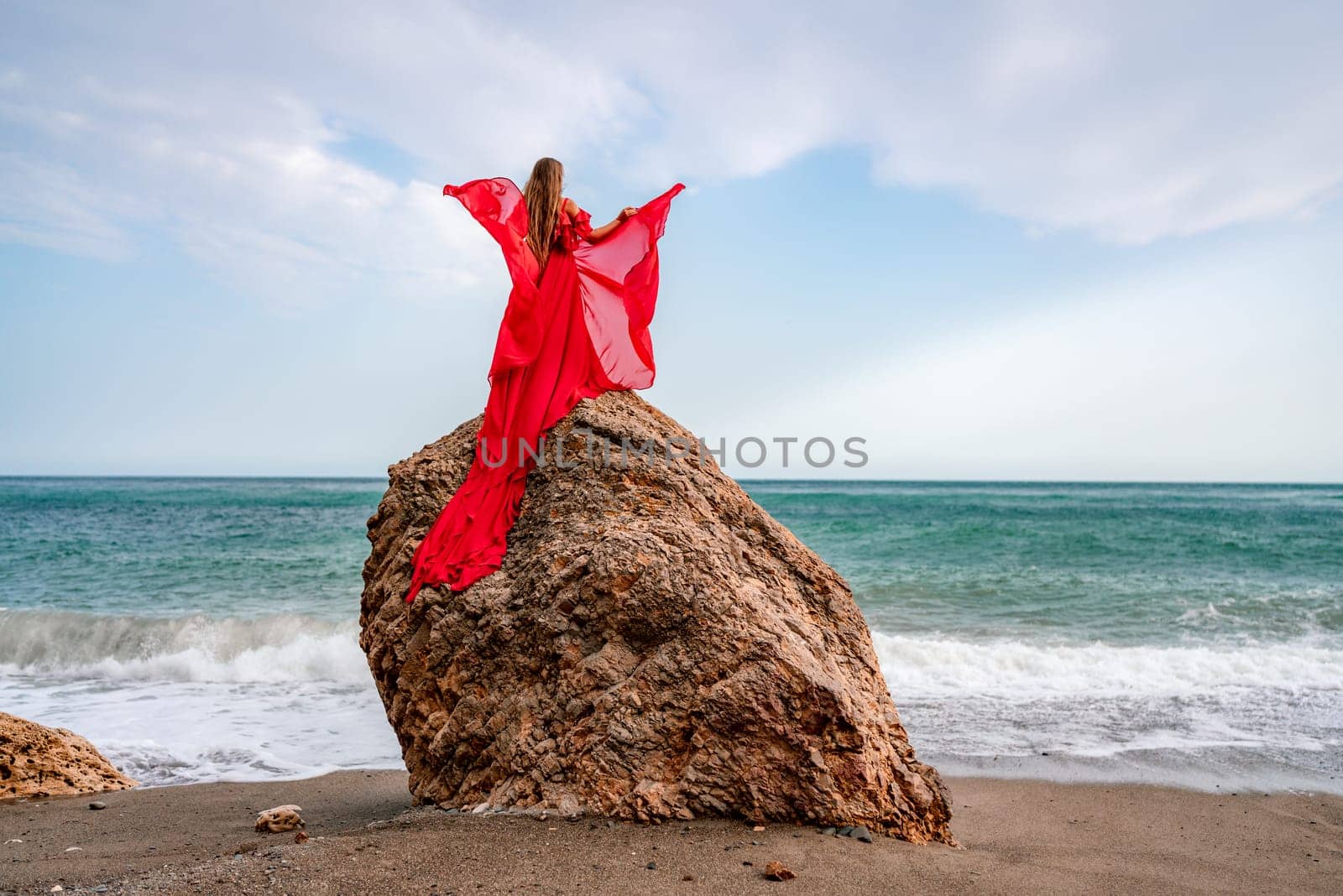 woman sea red dress. Woman with long hair on a sunny seashore in a red flowing dress, back view, silk fabric waving in the wind. Against the backdrop of the blue sky and mountains on the seashore