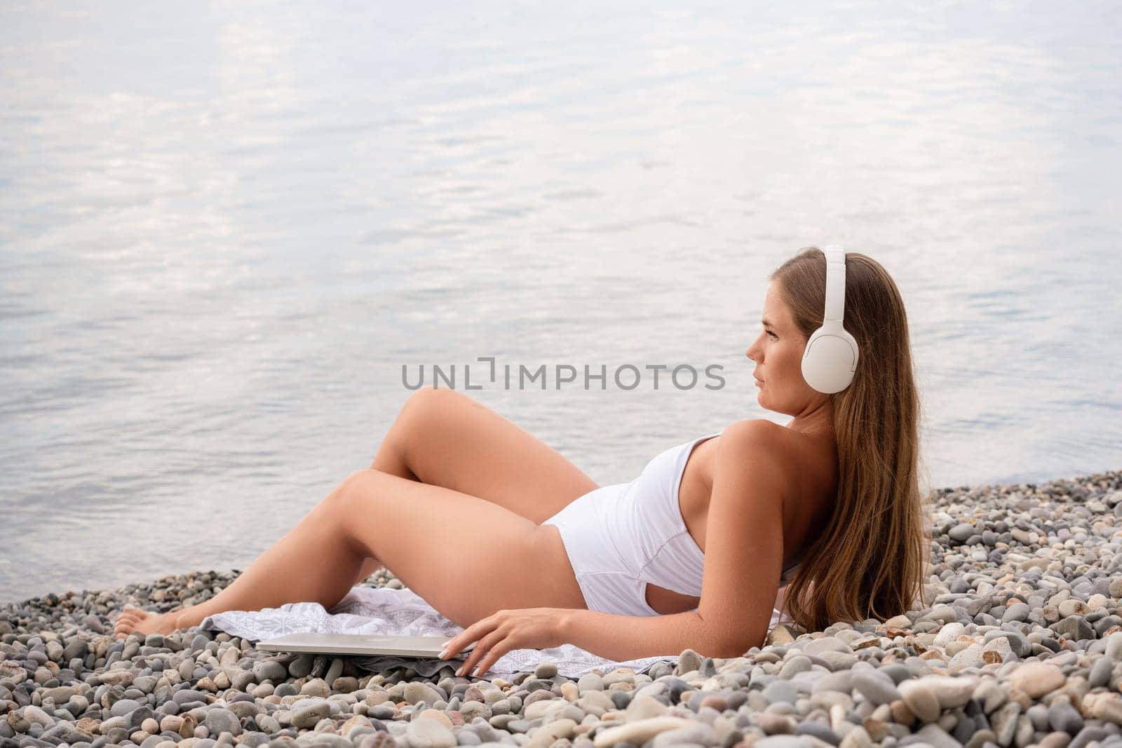 A woman is laying on the beach with a laptop and headphones on. She is wearing a white bikini and she is relaxing