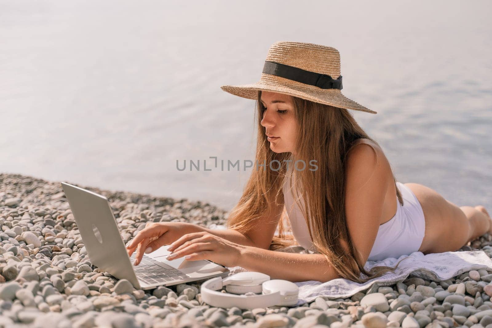 A woman is laying on the beach with a laptop on her lap. She is wearing a straw hat and a white bikini top. Concept of relaxation and leisure