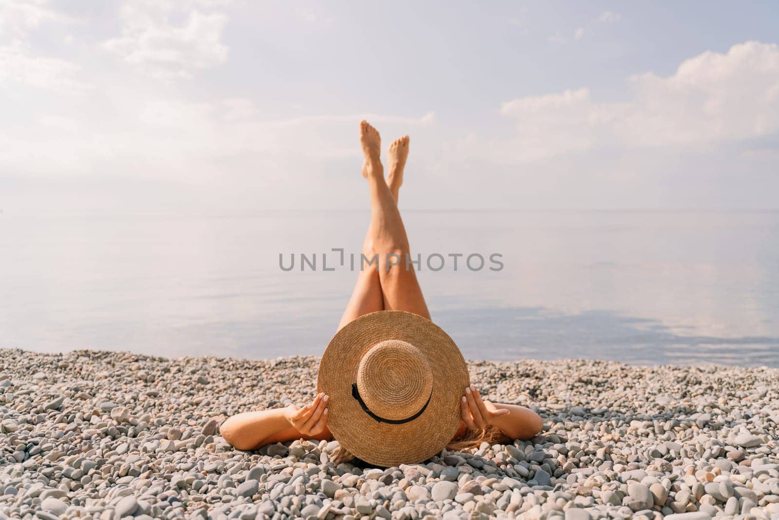 A woman is laying on the beach with her legs crossed and a straw hat on her head. The beach is rocky and the sky is cloudy