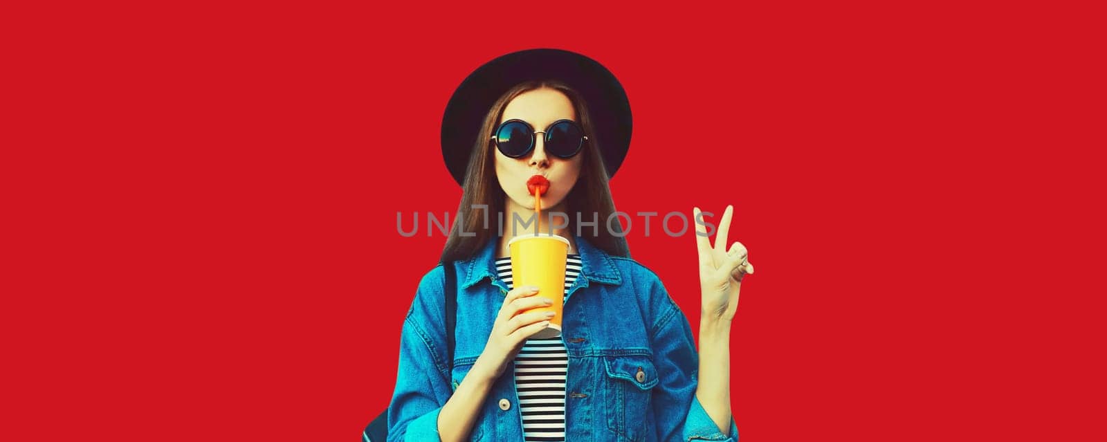 Stylish young woman drinking juice wearing black round hat, jean jacket on red background, blank copy space for advertising text