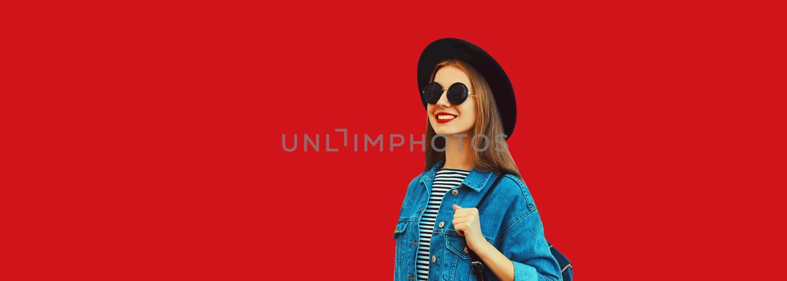 Portrait of happy smiling young woman looking away wearing black round hat, jean jacket, backpack on red background, blank copy space for advertising text