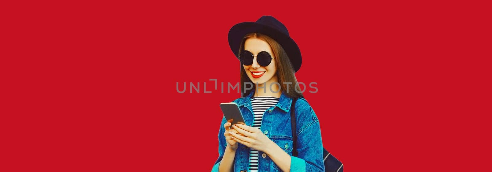 Summer portrait of happy traveler young woman 20s with mobile phone looking at device in black hat with backpack on red studio background