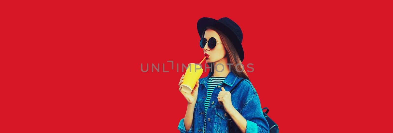 Stylish young woman drinking juice wearing black round hat, jean jacket on red background by Rohappy