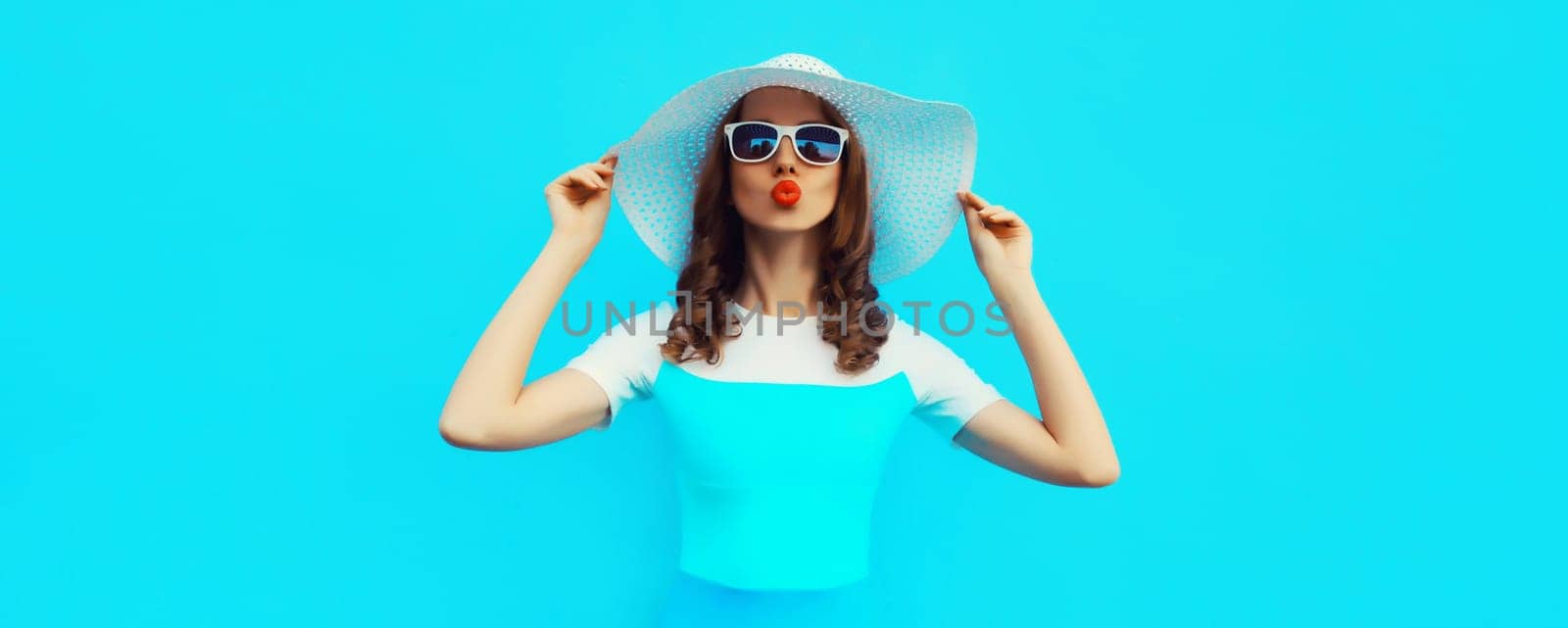 Portrait of beautiful young woman posing blowing kiss wearing summer straw hat and dress on blue background