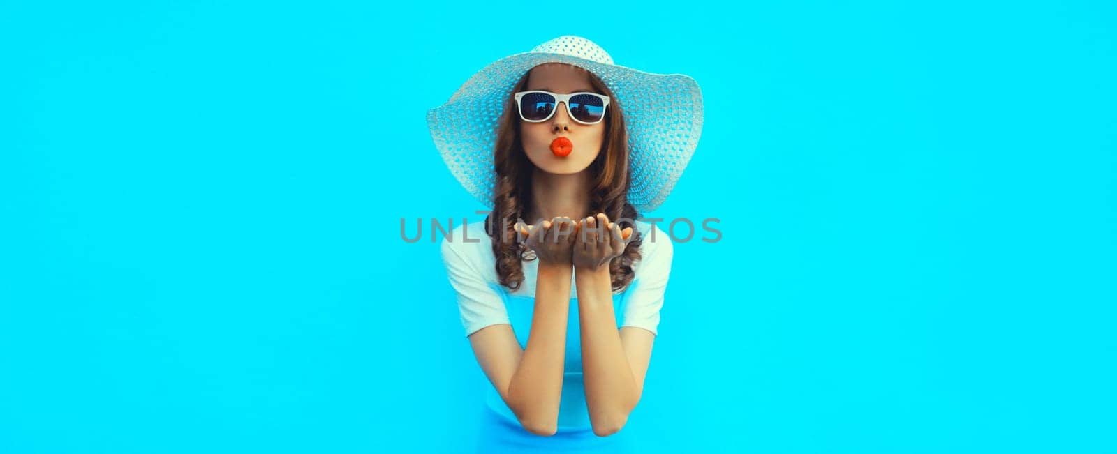 Portrait of beautiful young woman posing blowing kiss wearing summer straw hat and dress on blue background