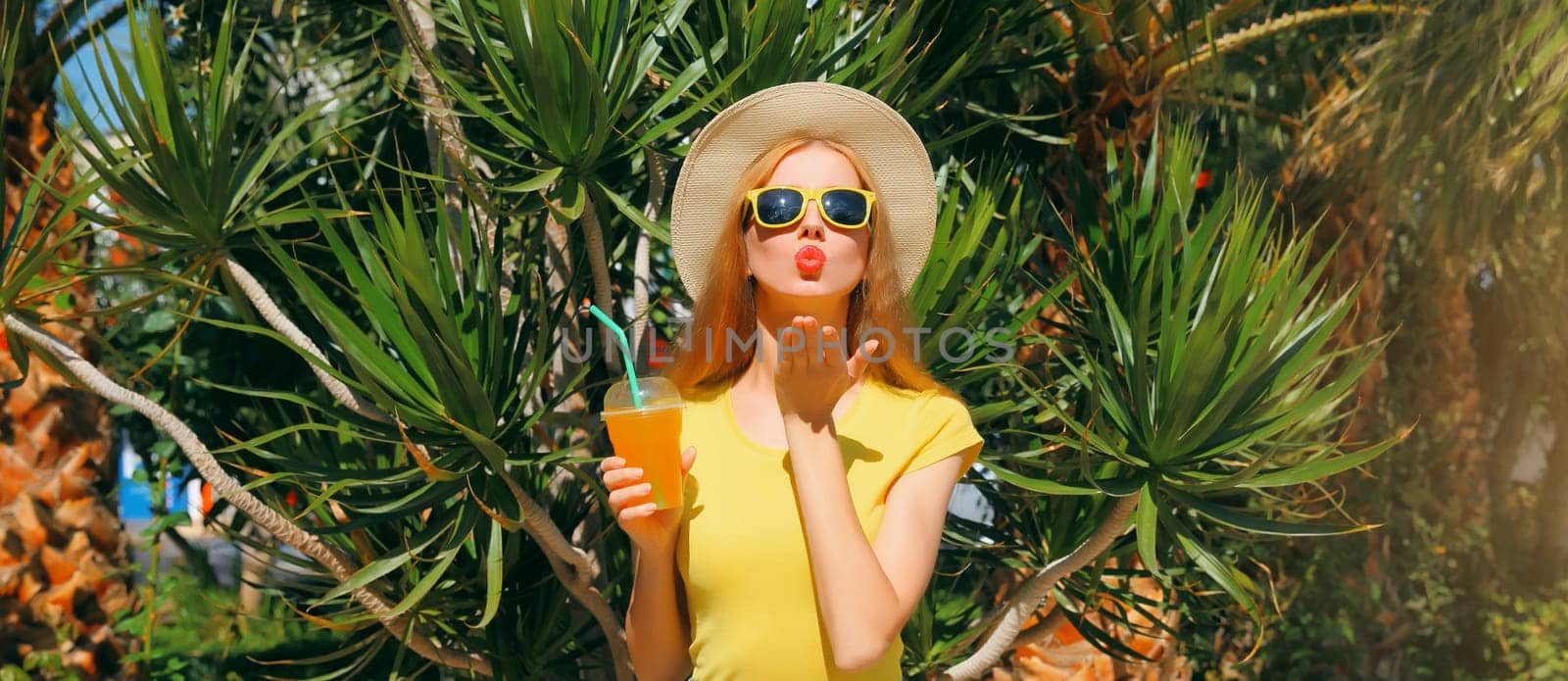 Summer sunny portrait of happy young woman drinking fresh juice wearing straw hat blowing a kiss on the beach with palm tree background