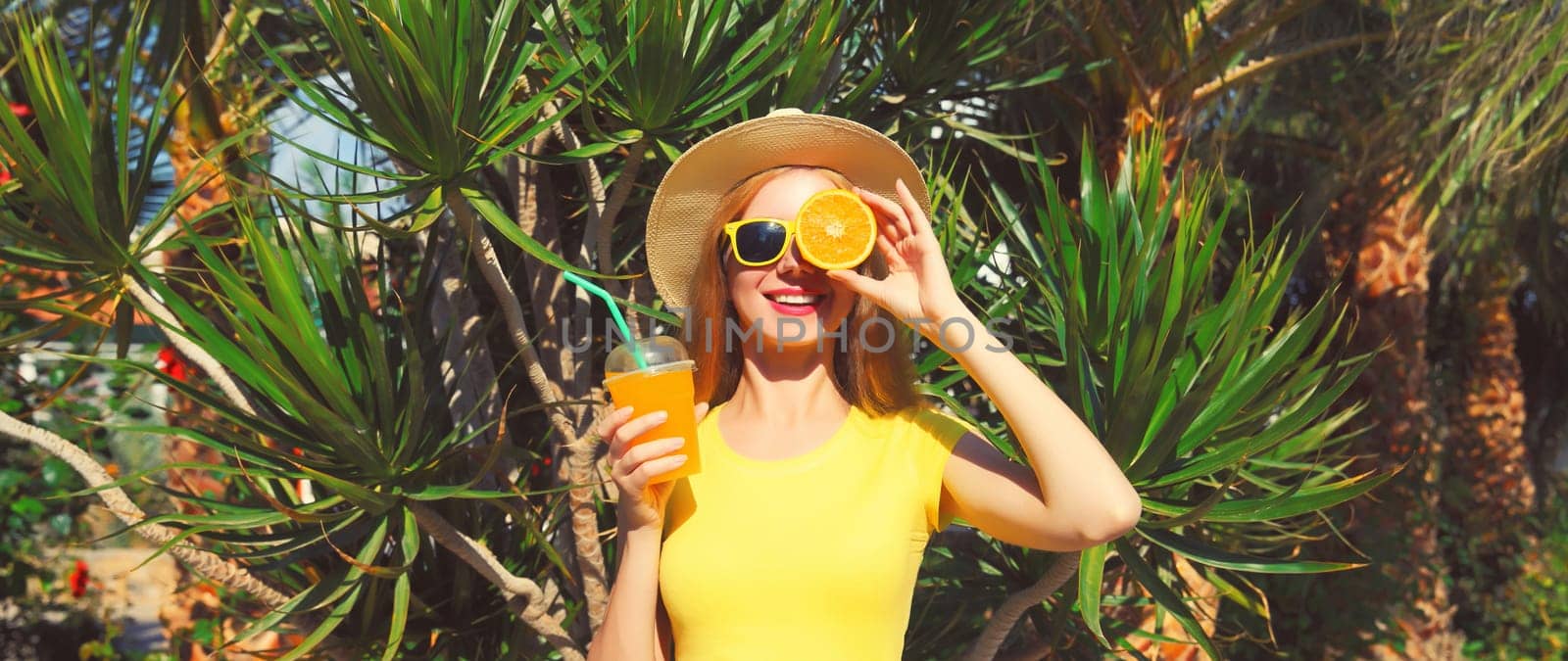 Summer portrait of happy smiling young woman drinking fresh juice with slice of orange fruit wearing straw hat on the beach with palm tree background
