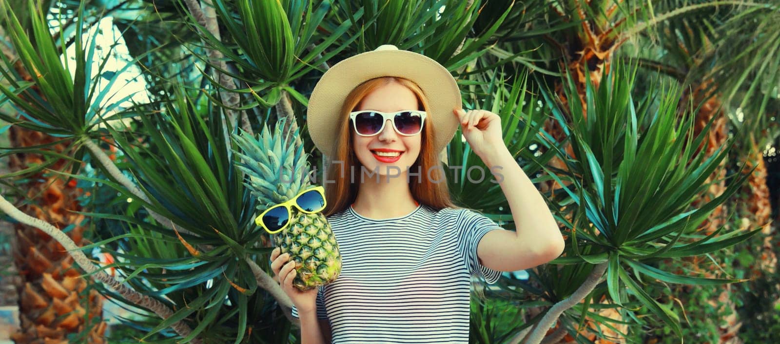 Summer portrait of happy smiling young woman with pineapple fruits wearing sunglasses, straw hat posing on palm tree background