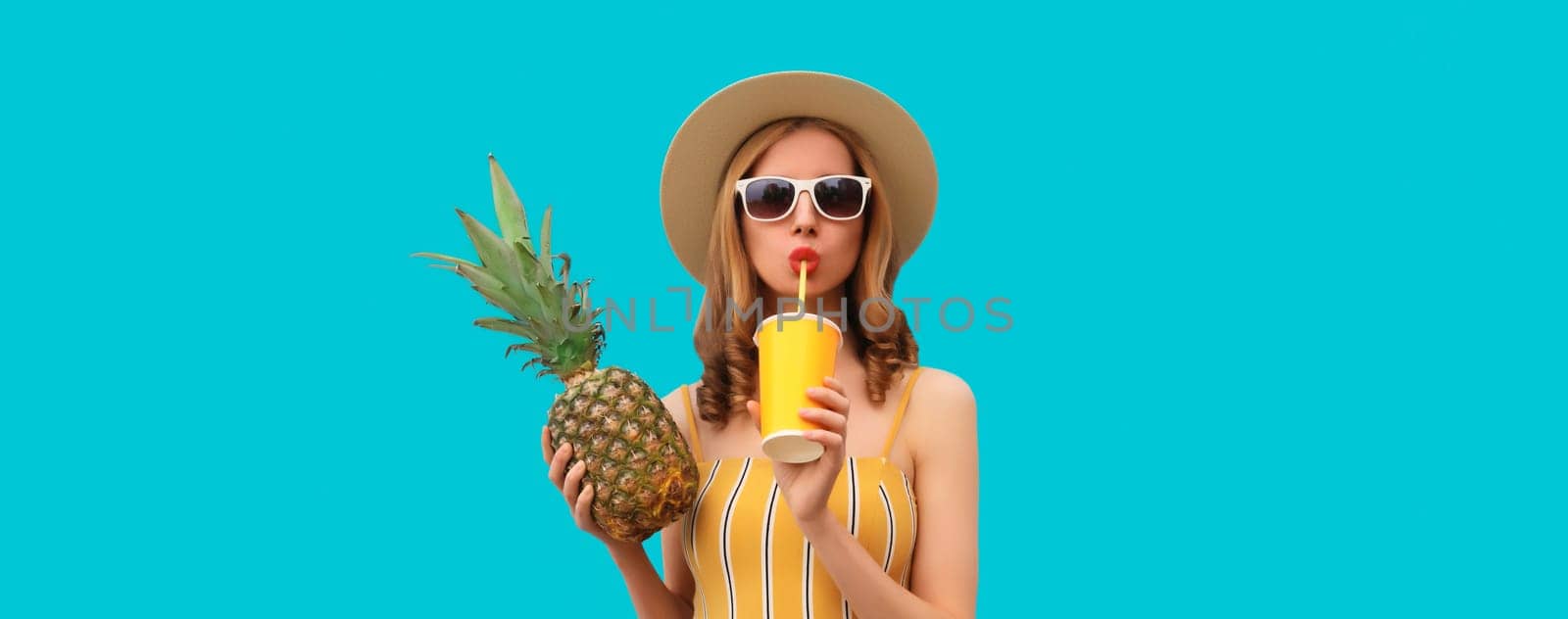 Summer bright portrait of stylish young woman drinking fresh juice holding pineapple fruit in straw hat, sunglasses on colorful blue studio background