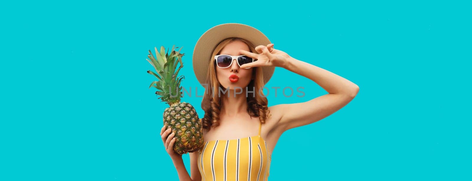 Summer portrait of beautiful young woman blowing a kiss with pineapple wearing straw hat, glasses posing on blue studio background