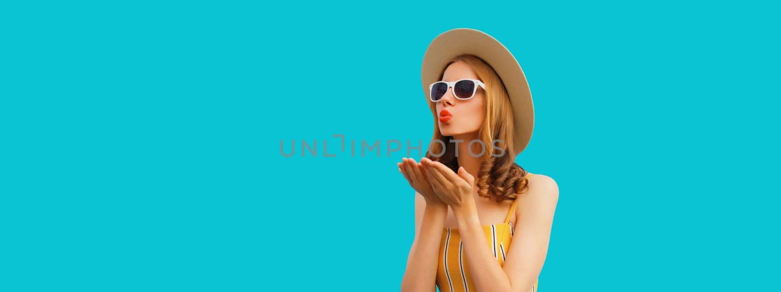 Portrait of beautiful stylish young woman blowing kiss in summer hat, glasses posing on blue background