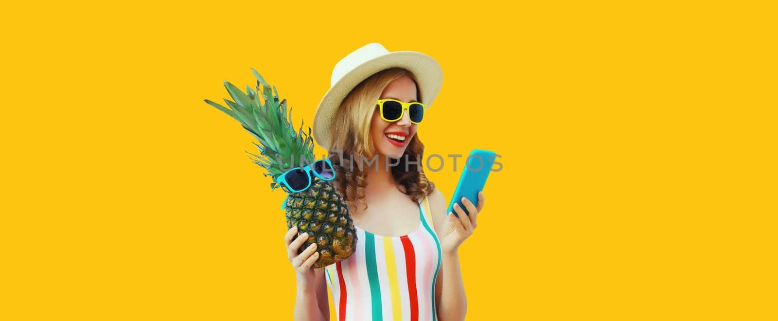 Summer portrait of traveler happy young woman with mobile phone holding fresh pineapple fruit looking at device in casual straw hat, sunglasses on bright yellow studio background