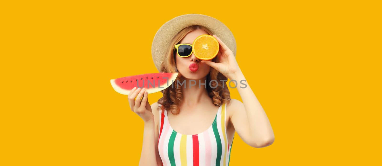 Summer portrait of happy young woman with fresh juicy fruits, slice of watermelon wearing straw hat, glasses and blowing kiss on bright yellow background