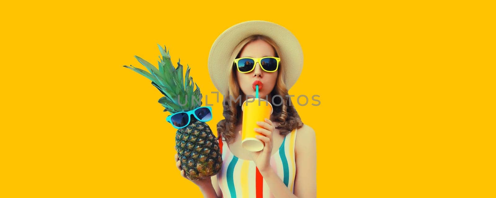 Summer bright portrait of stylish young woman drinking fresh juice holding pineapple fruit in straw hat, sunglasses on colorful yellow studio background