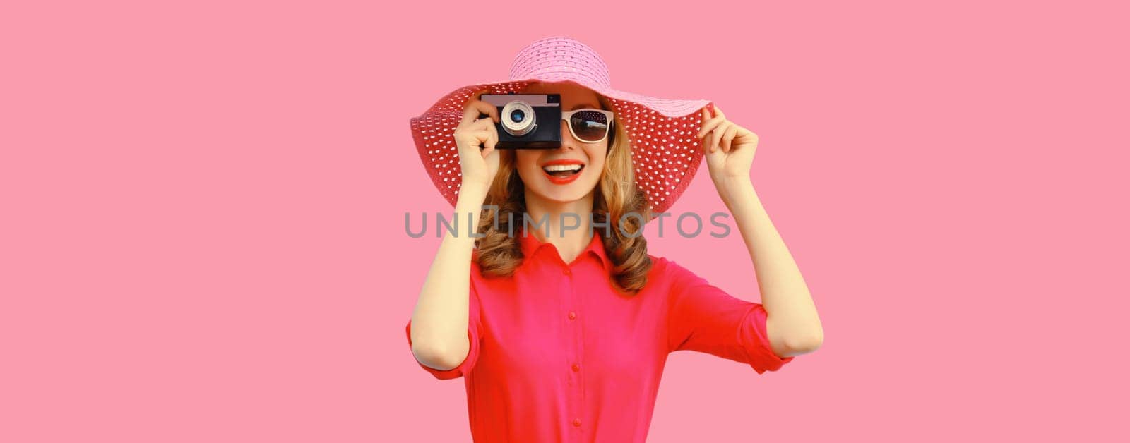 Summer portrait of happy smiling young woman photographer with film camera wearing straw hat, pink dress, sunglasses on background