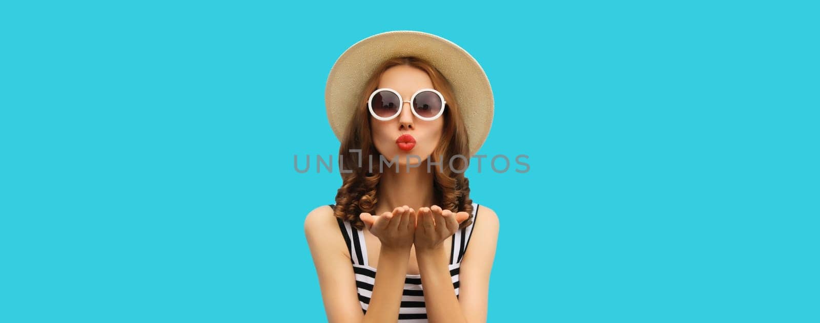 Portrait of beautiful young woman posing blowing kiss wearing summer straw hat and dress on blue background