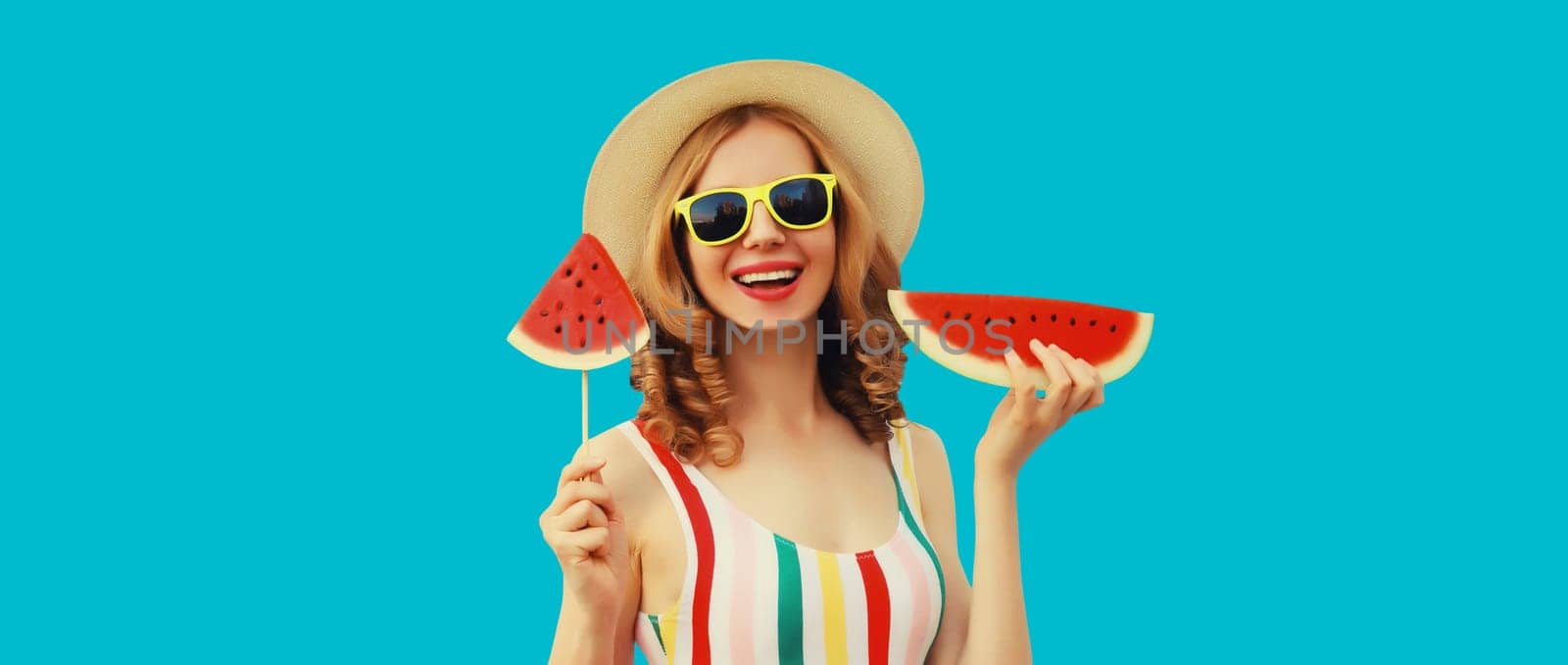 Summer portrait of happy young woman with lollipop or ice cream shaped slice of watermelon wearing straw hat on blue studio background