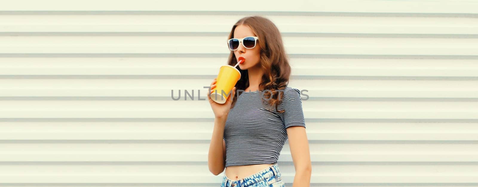 Stylish young woman drinking fresh juice wearing glasses looking away on white wall background